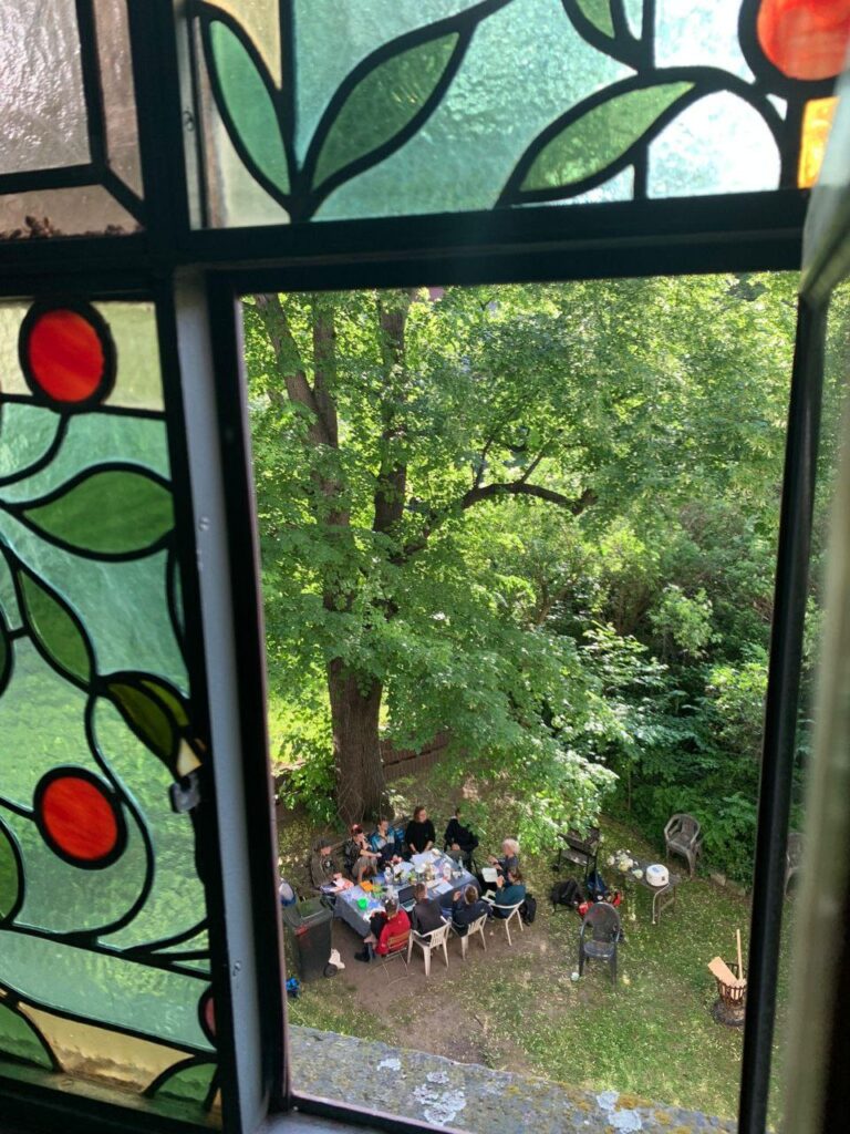 Der Blick durch ein buntverglastes Fenster fällt auf die Gruppe. Sie arbeitet im grünen Garten